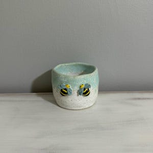 May include: A small, handmade ceramic pot with a speckled white base and a light blue rim. Two yellow and black bee figurines are attached to the front. The pot is likely for holding small plants or trinkets.