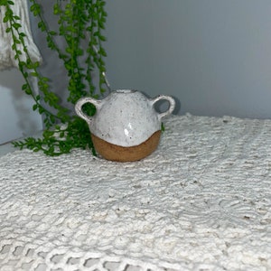 May include: A small white ceramic vase with two handles and a brown rim. The vase is sitting on a white crocheted surface.
