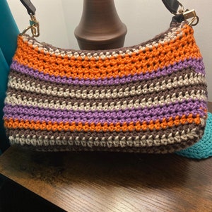 May include: A handmade crochet shoulder bag with a black strap and gold-coloured hardware. The bag features horizontal stripes in shades of orange, purple, brown, and cream. The bag is sitting on a wooden surface.