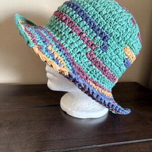 May include: A multi-coloured crocheted sun hat. The hat has a turquoise base with stripes of purple, blue, and yellow. The brim is slightly curved and has a woven texture. The hat is displayed on a white mannequin head.
