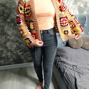 Può includere: Un cardigan all'uncinetto con una base marrone e quadrati colorati in rosso, giallo e bianco. Il cardigan ha una vestibilità ampia e un lungo davanti aperto.