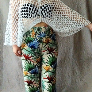 May include: A white crochet top with a loose, flowing fit. The top has a large, open weave and a tie at the neck. The model is wearing the top over a black bra and floral print trousers.