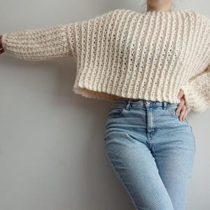 May include: A cream-colored, long-sleeved, cropped sweater featuring a vertical ribbed knit pattern with a wide, loose fit and a round neckline, paired with light blue denim jeans that have a classic button and zipper fly closure.