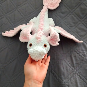 May include: A handmade white and pink crochet dragon plush toy with orange eyes. The dragon has a long tail and wings, and is laying on a gray quilted surface. The dragon is made of soft, plush yarn.