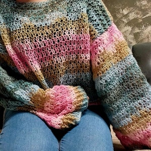May include: A crocheted sweater with a striped pattern in shades of blue, pink, and brown. The sweater has a loose fit and a wide neckline.