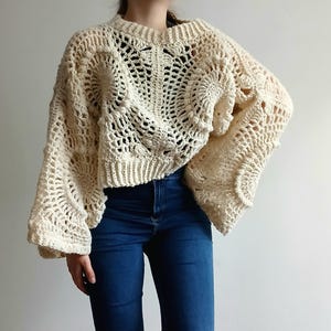 May include: An off-white, long-sleeved crochet sweater featuring a complex, circular openwork pattern throughout the body and sleeves. The garment has a ribbed crew neckline and ribbed cuffs, with a cropped length that sits above blue denim jeans.