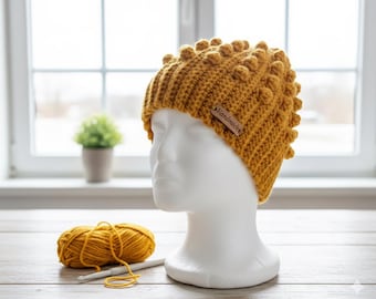 May include: Mustard yellow crochet beanie with a textured bobble stitch design. The hat is displayed on a white mannequin head. A small brown tag reads "Handmade". A ball of yellow yarn and a crochet hook are in the foreground.