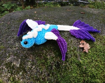 May include: A handmade, plush creature with a blue body, white accents, and purple wings and tail. The toy has large, black eyes and is lying on a moss-covered rock. The plushie is a whimsical, soft toy.