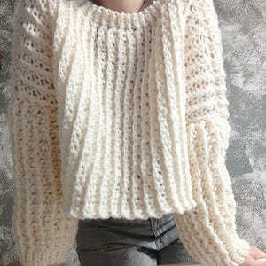 May include: Cream-colored, hand-crocheted sweater with a ribbed design. The sweater features long sleeves and a crew neck. The yarn appears thick and soft, with a textured stitch pattern. The garment is a cozy, handmade piece of apparel.