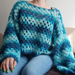 May include: A long-sleeved crochet sweater featuring a loose, open-knit granny square pattern in various shades of blue, including navy, royal blue, and light turquoise. The garment has a wide, relaxed fit with a round neckline and long sleeves.