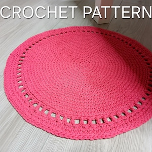 May include: A round, bright pink crochet rug with a decorative border. The rug is made of thick yarn and has a textured surface. The words "CROCHET PATTERN" are in white text at the top of the image.