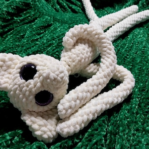 Może przedstawiać: Biała pluszowa zabawka wykonana na szydełku z dużymi fioletowymi oczami. Zabawka leży na zielonej pluszowej powierzchni.