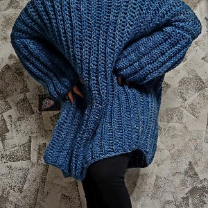 Può includere: Un maglione all'uncinetto blu, grosso e oversize. Il maglione ha uno scollo ampio e a coste e maniche lunghe. Il punto all'uncinetto crea un motivo a trama aperta e strutturata. L'indumento è indossato con leggings neri.