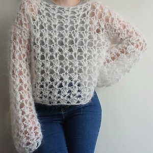 May include: A white, long-sleeved, loose-knit sweater featuring a semi-sheer, open-work pattern with a fuzzy, mohair-like texture. The garment has a wide, rounded neckline and a cropped silhouette, showcasing a delicate, airy construction.