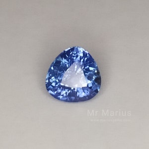 Op de afbeelding: Een driehoekige blauwe saffier edelsteen met een facetgeslepen vorm. De edelsteen is geplaatst tegen een witte achtergrond.