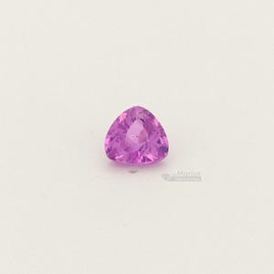 May include: A triangular, faceted, pink gemstone. The gem has a polished surface and a vibrant, light pink color. The gemstone is set against a white background. The image is well-lit, highlighting the gem's clarity and shape.