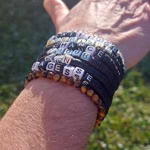 Peut inclure: Un empilement de sept bracelets composés de perles disques noires, de perles en œil de tigre marron et de perles carrées blanches avec des lettres. Les perles forment les mots GRATITUDE, CHANCE, AMOUR et SAGESSE. Les bracelets sont superposés.