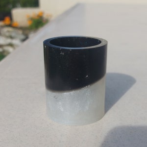 Peut inclure: Un récipient cylindrique, divisé en deux parties, noire et blanche nacrée. La partie supérieure est noire, tandis que la partie inférieure est blanche nacrée. Le récipient a une finition brillante et semble être en résine.