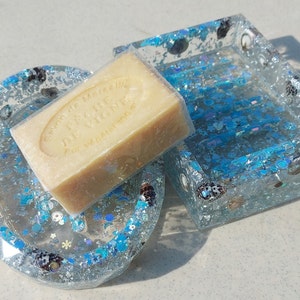 Peut inclure: Deux porte-savons transparents avec des paillettes bleues et des coquillages. Une barre de savon avec le texte "Savon de Marseille Pêche Divine" est posée sur l'un des porte-savons.