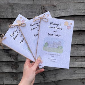 May include: Three folded wedding ceremony programmes with a watercolour illustration of a church and autumn leaves. The programmes are tied together with twine and have the text "Marriage of Hannah Holsey and Elliott Jackson" and "Saturday 12th November 2022 11am All Saints Church East Pennard".