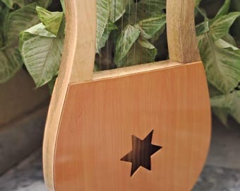 May include: A wooden lyre musical instrument featuring a light brown finish, a U-shaped frame, and a central star-shaped sound hole. The instrument includes multiple thin vertical strings stretched across the open frame and a smooth, polished surface.