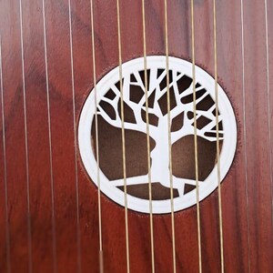 May include: Close-up of a stringed instrument with a dark reddish-brown wooden body. The instrument features a white, circular sound hole with a tree design. Numerous strings run across the body.