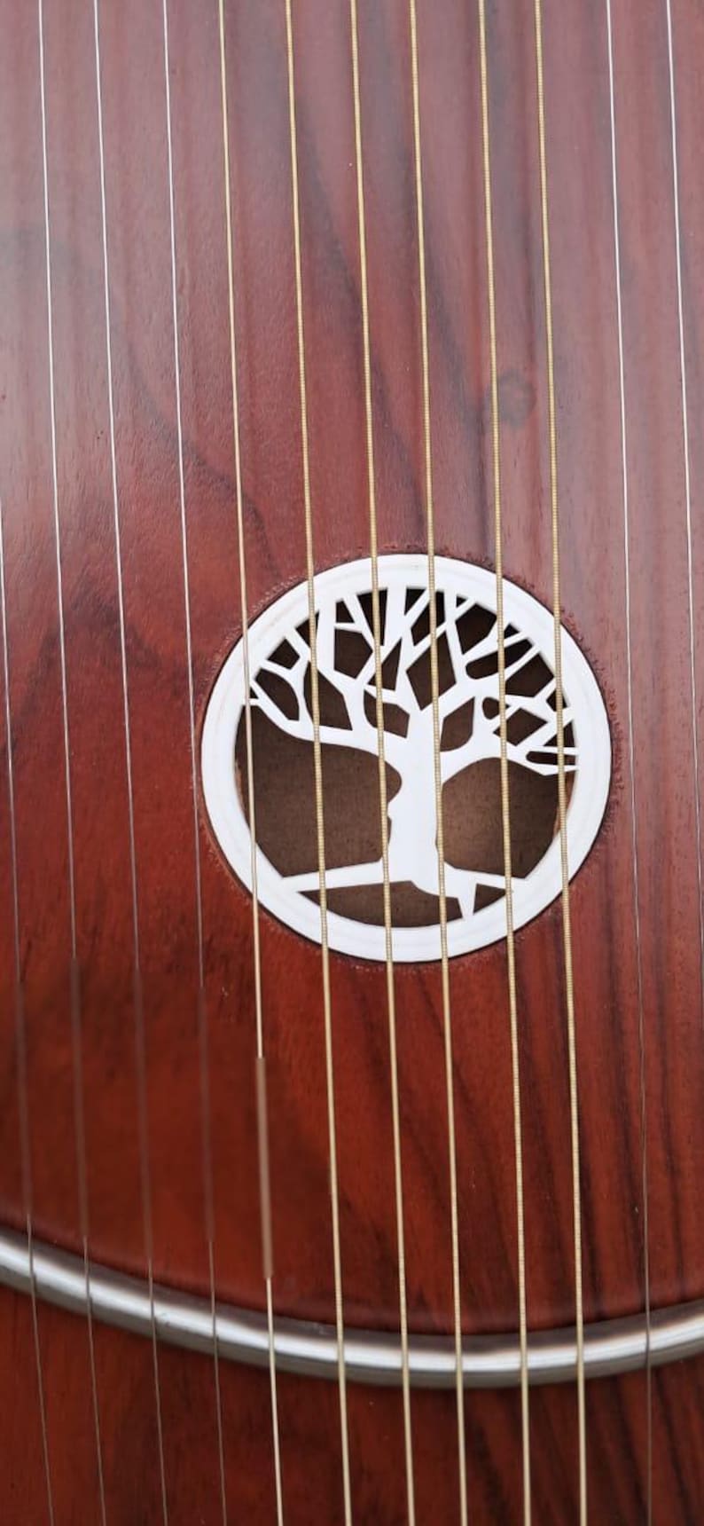 May include: Close-up of a stringed instrument with a dark reddish-brown wooden body. The instrument features a white, circular sound hole with a tree design. Numerous strings run across the body.