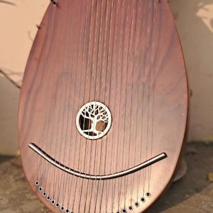 Puede incluir: Instrumento musical de madera con numerosas cuerdas, con forma de lágrima y orificio de sonido curvo. Presenta un diseño de árbol. Las cuerdas se extienden por el cuerpo del instrumento.