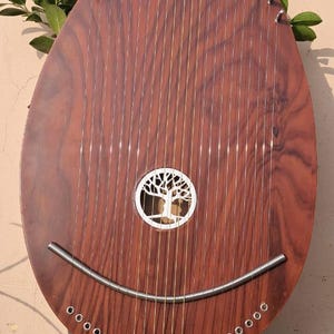 May include: A brown wooden musical instrument with numerous strings, a tree design, and a curved metal piece. The instrument is set in a black pot, with green leaves in the background. The instrument's shape is oval.