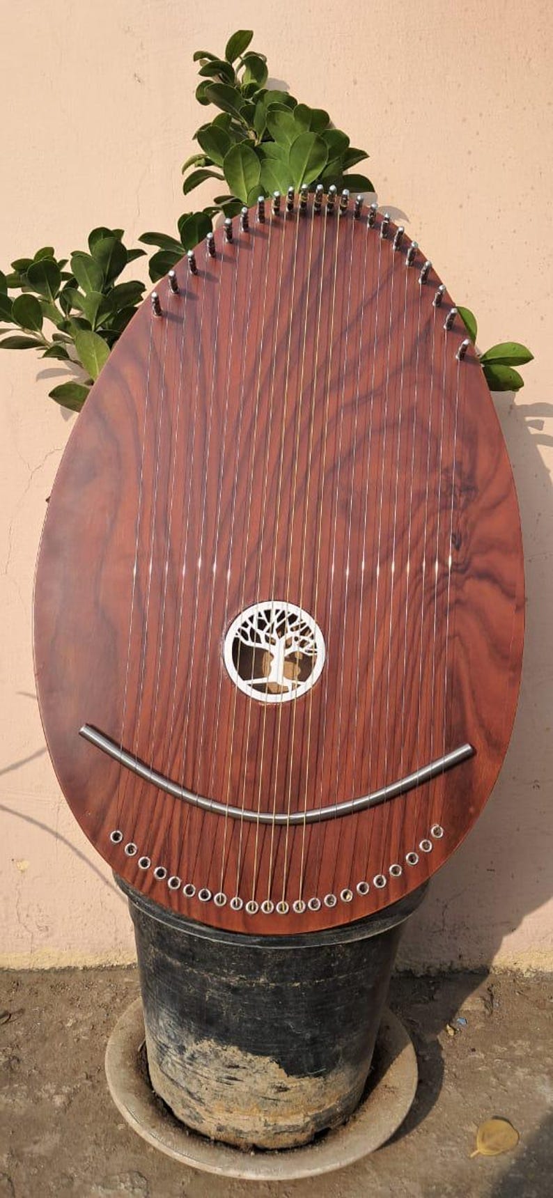 May include: A brown wooden musical instrument with numerous strings, a tree design, and a curved metal piece. The instrument is set in a black pot, with green leaves in the background. The instrument's shape is oval.