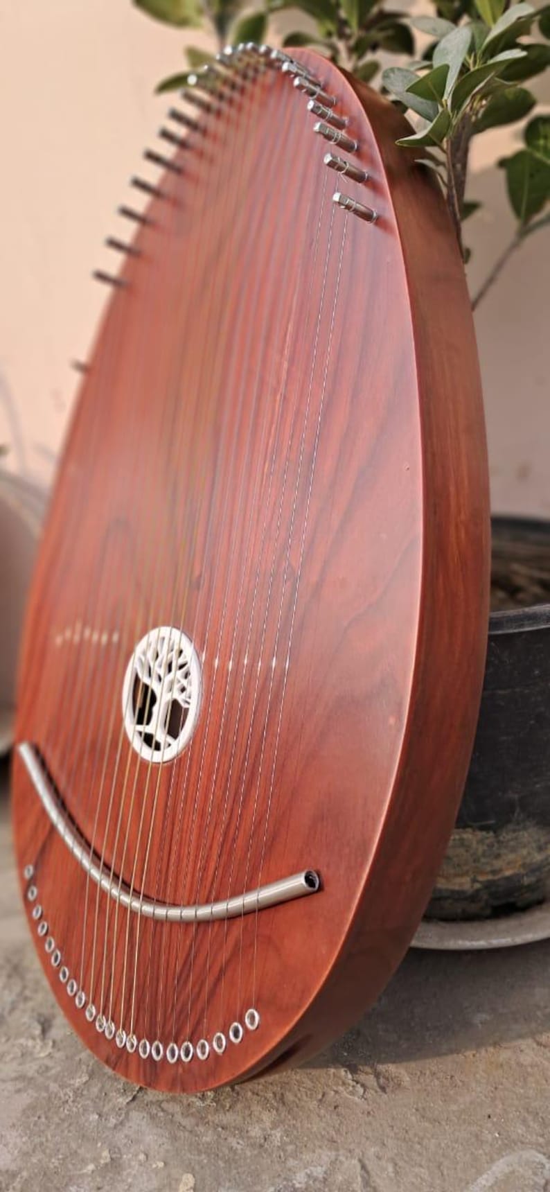 May include: A wooden instrument with a teardrop shape, featuring numerous strings and a sound hole with a tree design. The instrument has a rich, reddish-brown finish and a curved metal piece at the bottom.