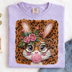 Op de afbeelding: Lichtpaars T-shirt met een gedetailleerde illustratie van een konijn met een bril en een bloemenkrans, die een kauwgomblaas blaast. Het konijn is geplaatst tegen een gebreide achtergrond.