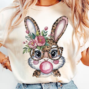 Può includere: T-shirt color crema con un design a coniglio stravagante. Il coniglio ha una corona floreale, occhiali e una bolla di gomma da masticare. Il design incorpora stampe leopardate, a quadri e floreali. Una maglietta grafica divertente e colorata.