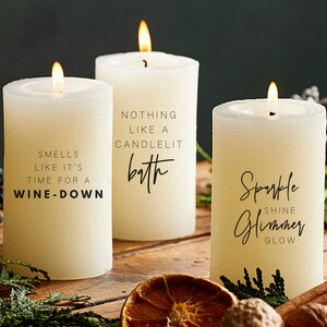 May include: Three white pillar candles with black text. The candles are on a wooden surface with cinnamon sticks, nuts, and a dried orange slice. The text on the candles reads: "Smells like it's time for a wine-down", "Nothing like a candlelit bath", and "Sparkle Shine Glimmer Glow".