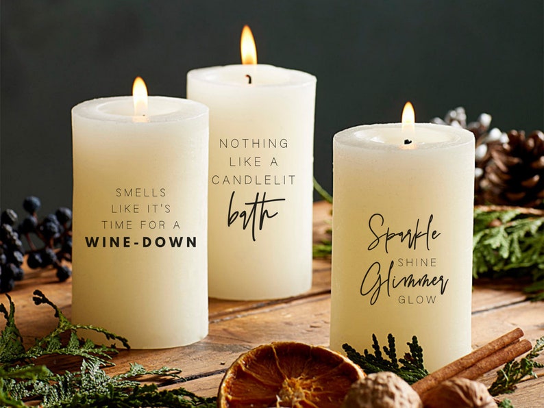 May include: Three white pillar candles with black text. The candles are on a wooden surface with cinnamon sticks, nuts, and a dried orange slice. The text on the candles reads: "Smells like it's time for a wine-down", "Nothing like a candlelit bath", and "Sparkle Shine Glimmer Glow".