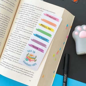 May include: A colourful bookmark featuring a stack of books, a cup of coffee, and the text "Solo un Capitulo Mas". The bookmark also has five colourful stripes with the text "Romantico", "Frases Fav", "Plot Twist", "Triste", and "Solo un Capitulo Mas".
