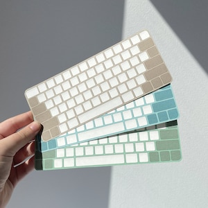 Può includere: Tre adesivi per tastiera in diversi colori: beige, blu e verde. Ogni adesivo ha un layout di tastiera con tasti bianchi e neri.