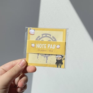 Puede incluir: Bloc de notas amarillo con una ilustración en blanco y negro de un personaje con un sombrero negro y una camisa blanca. El bloc de notas tiene una ilustración en blanco y negro de una calavera con huesos cruzados en la parte delantera. El bloc de notas dice "NOTE PAD 24 páginas / 80 gr" en la parte delantera.