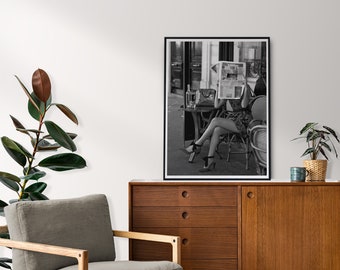 May include: A black and white framed wall art print depicting a person sitting at an outdoor cafe table, holding a newspaper in front of their face. The person wears a patterned dress and high-heeled shoes with ankle ties, seated on a wicker chair.