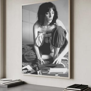 Puede incluir: Fotografía en blanco y negro enmarcada de una persona sentada cerca de una máquina de escribir antigua. La persona lleva pantalones oscuros y una parte superior clara. La fotografía se muestra sobre una superficie gris, con libros y un bloc de notas en primer plano.