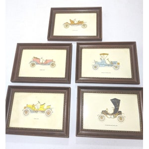 May include: Five framed vintage car illustrations in brown frames with gold trim. Each illustration depicts a different car model from the early 1900s. The cars are in various colours, including yellow, blue, red, and black.