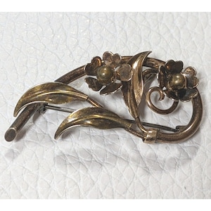 May include: A gold-toned brooch featuring two stylized flowers with green leaves. The brooch has a pin on the back for attaching to clothing.