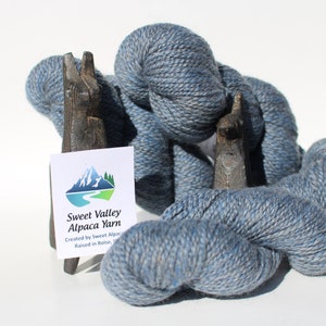 May include: Three skeins of blue alpaca yarn, wrapped around wooden llama figurines. The yarn is a light blue with a subtle fleck of white. A white tag with the text "Sweet Valley Alpaca Yarn" is visible in the center of the image.