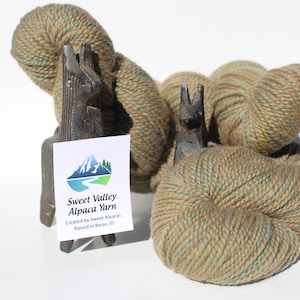 May include: Two skeins of alpaca yarn in a green and brown colorway, displayed on wooden llama figurines. The yarn is labeled "Sweet Valley Alpaca Yarn, Created by Sweet Alpacas, Raised in Boise, ID."