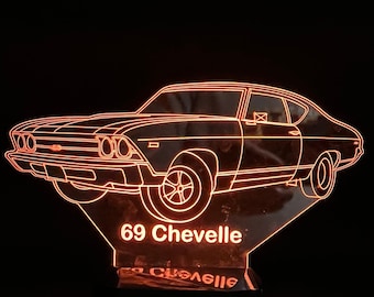May include: An illuminated acrylic sign featuring a 1969 Chevelle car outline. The car is depicted in orange light, with the text "69 Chevelle" at the base. The sign is mounted on a black base, creating a striking contrast against the dark background.