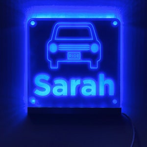 May include: A blue illuminated sign with a car silhouette and the name "Sarah" in white lettering. The car has the word "RIDE" on the license plate.