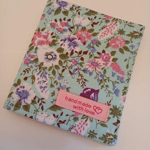 May include: A rectangular fabric wallet featuring a light blue background with a floral pattern in shades of pink, purple, and dark green. A small, rectangular pink fabric label is stitched to the front with the text "hand made with love" and a heart icon.