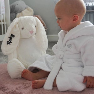 May include: A white plush bunny with long ears and a pink nose. The bunny has the name "Ruby" embroidered on one ear. A baby is wearing a white hooded robe with an embroidered elephant on the front. The baby is sitting on a pink rug and looking at the bunny.