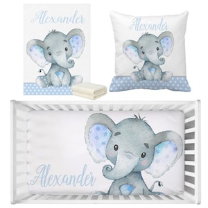 Puede incluir: Un juego de ropa de cama para cuna de bebé con un diseño de elefante azul y blanco. El juego incluye una sábana bajera, una funda de almohada y una manta. El elefante sostiene un corazón azul y tiene lunares azules en las orejas. El nombre "Alexander" está escrito en cursiva azul en la sábana bajera y la funda de almohada.