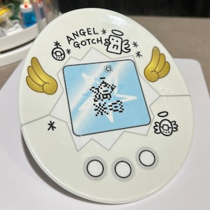 Pu&ograve; includere: Videogioco elettronico portatile bianco con la scritta "ANGEL GOTCH" e una grafica pixelata sullo schermo. Il dispositivo presenta ali d'angelo color oro e una piccola illustrazione di angelo. Tre pulsanti circolari si trovano in basso.